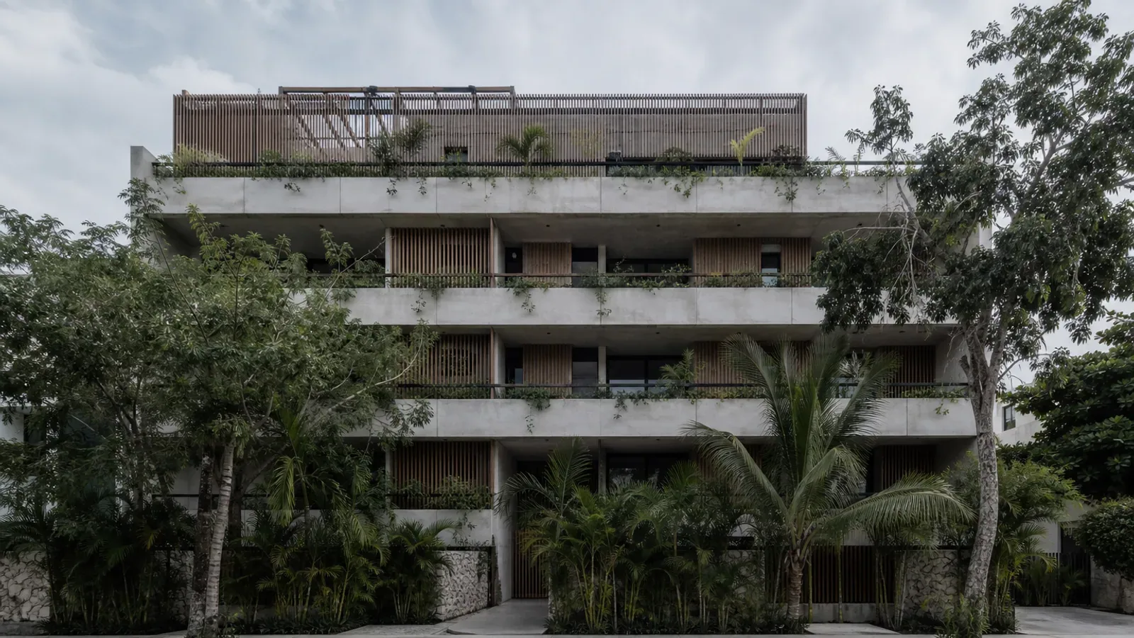 Edificio de departamentos de baja altura en la Riviera Maya visto desde la calle: balcones escalonados, celosías de madera y vegetación madura.